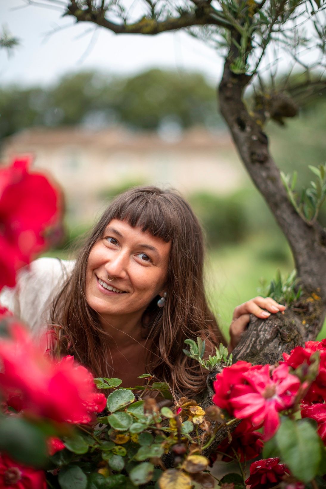 Smiling brunette woman lying among vibrant red flowers beside a tree in a garden.