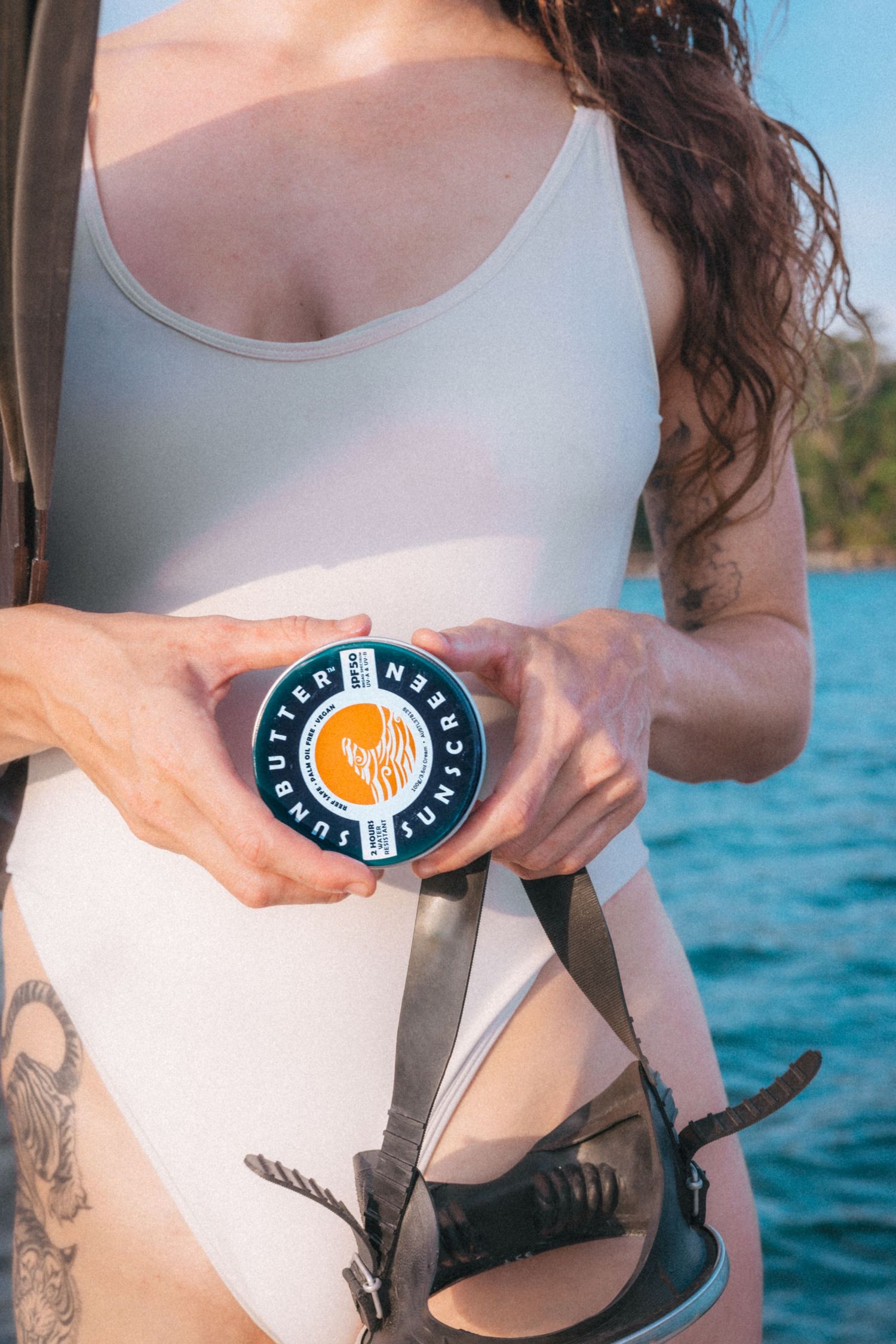 Woman in white swimsuit holding a round tin of SunButter sunscreen while standing near blue ocean water.