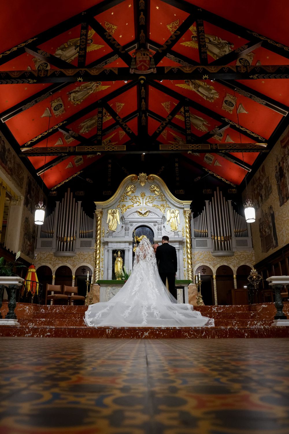 Wedding couple standing before ornate church altar with dramatic red ceiling and pipe organ.