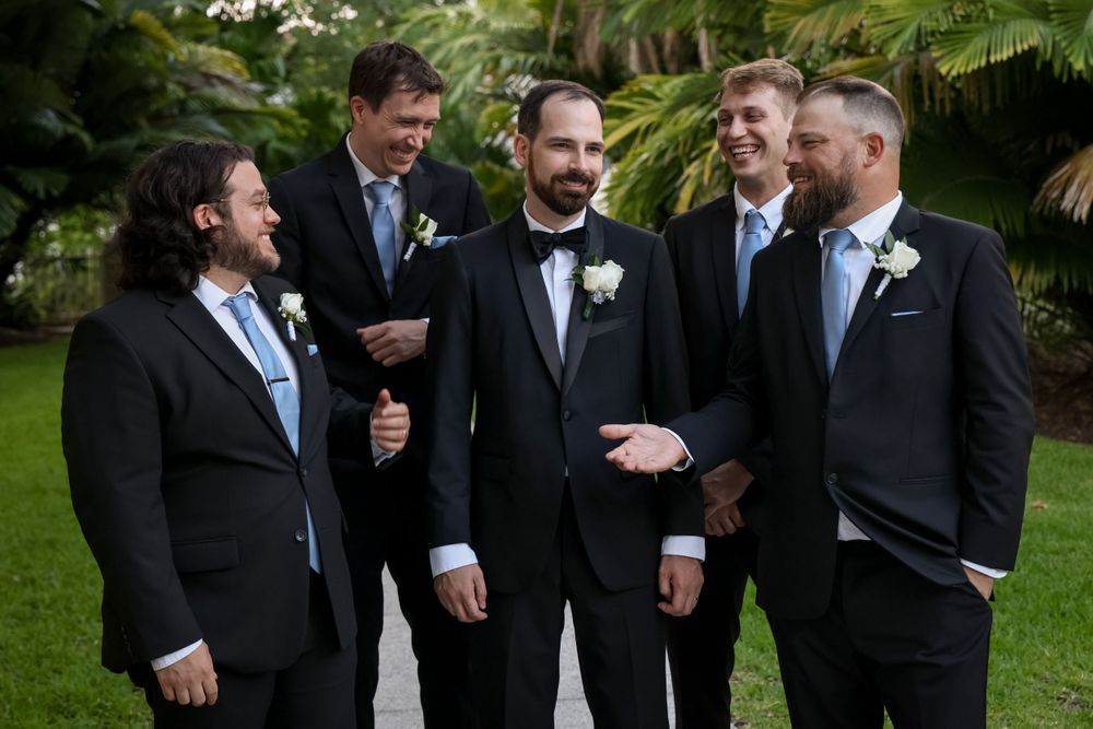 Groomsmen in formal black suits and blue ties share a candid laughing moment in a garden setting.