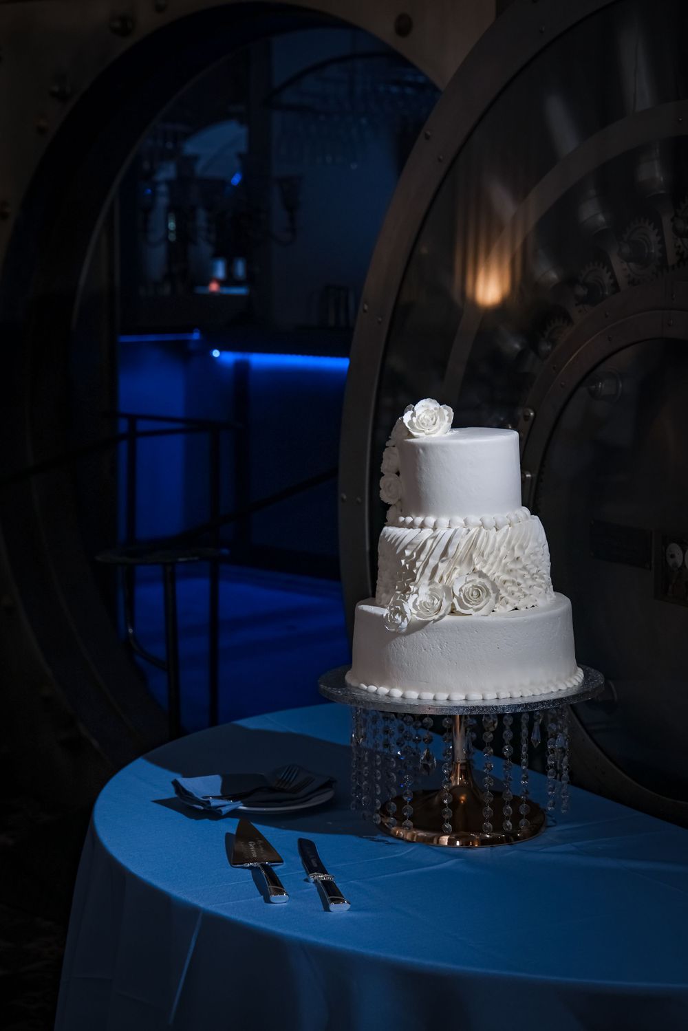 Elegant three-tier white wedding cake on crystal stand illuminated by moody blue lighting.
