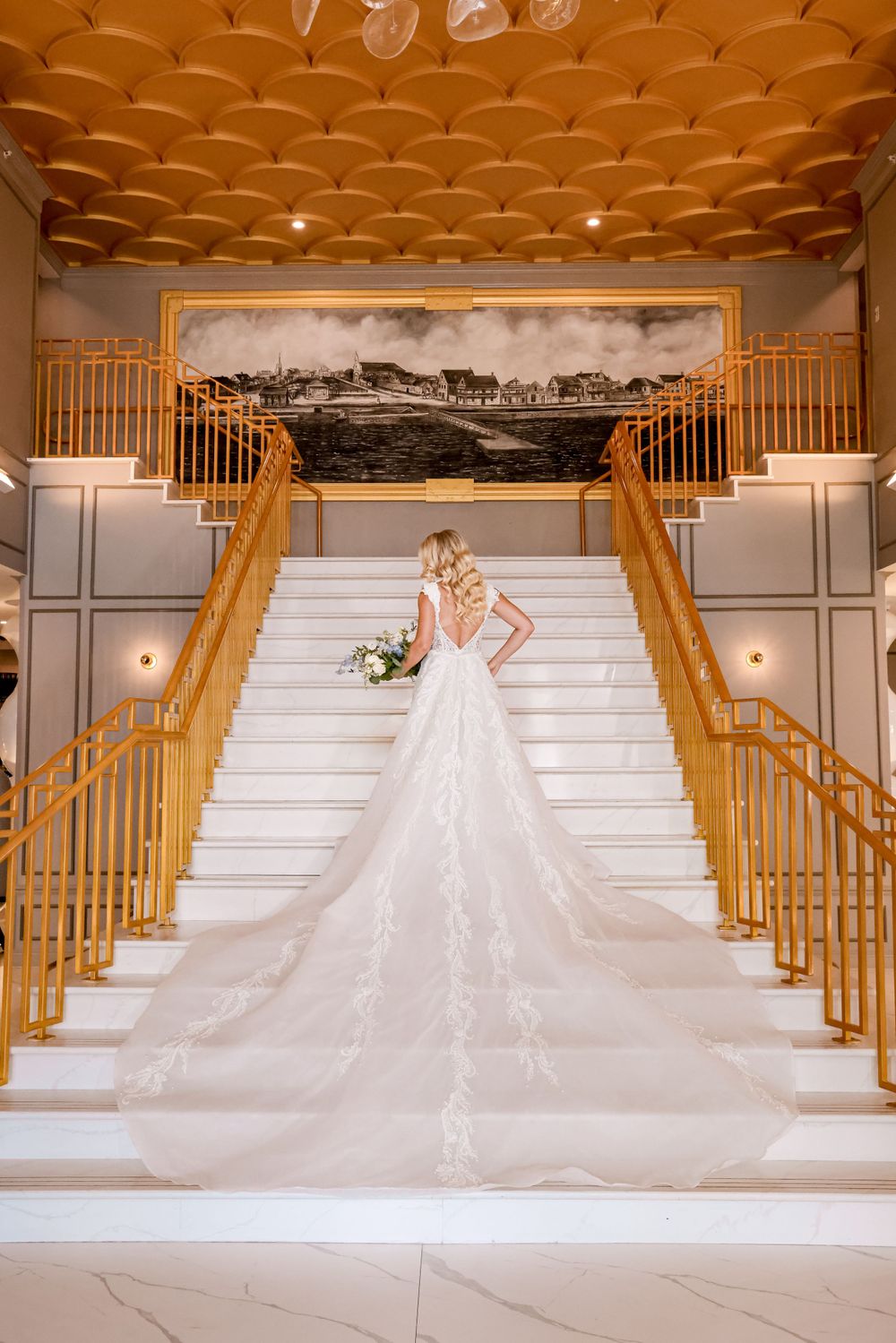 Wedding dress with long train displayed on grand white staircase with gold railings and decorative ceiling.