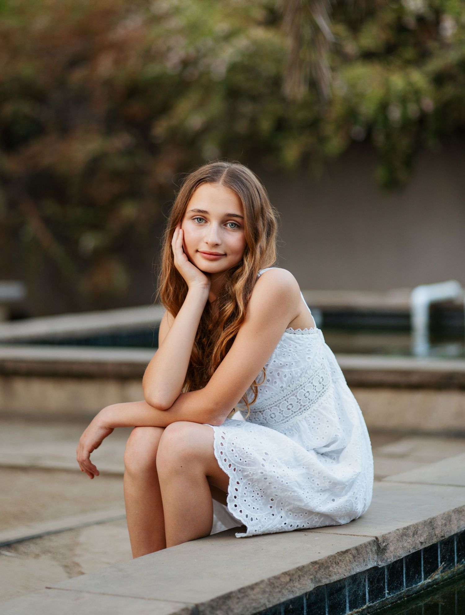 Young woman in white lace dress sitting thoughtfully by poolside at dusk.