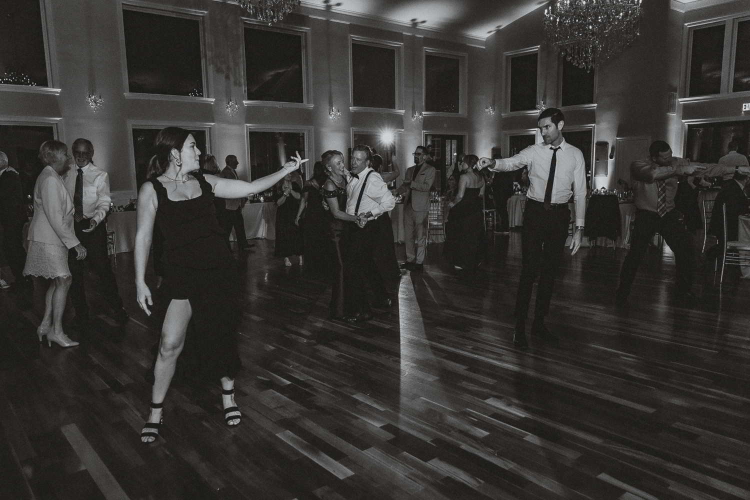 Black and white photo of wedding guests dancing on a polished wooden floor at a reception.