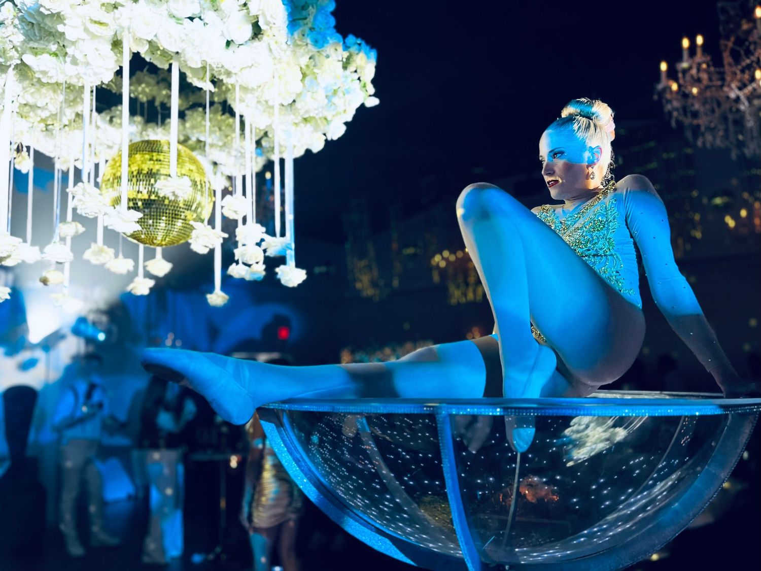 A performer in blue costume poses dramatically on a glass sphere under hanging floral decorations.
