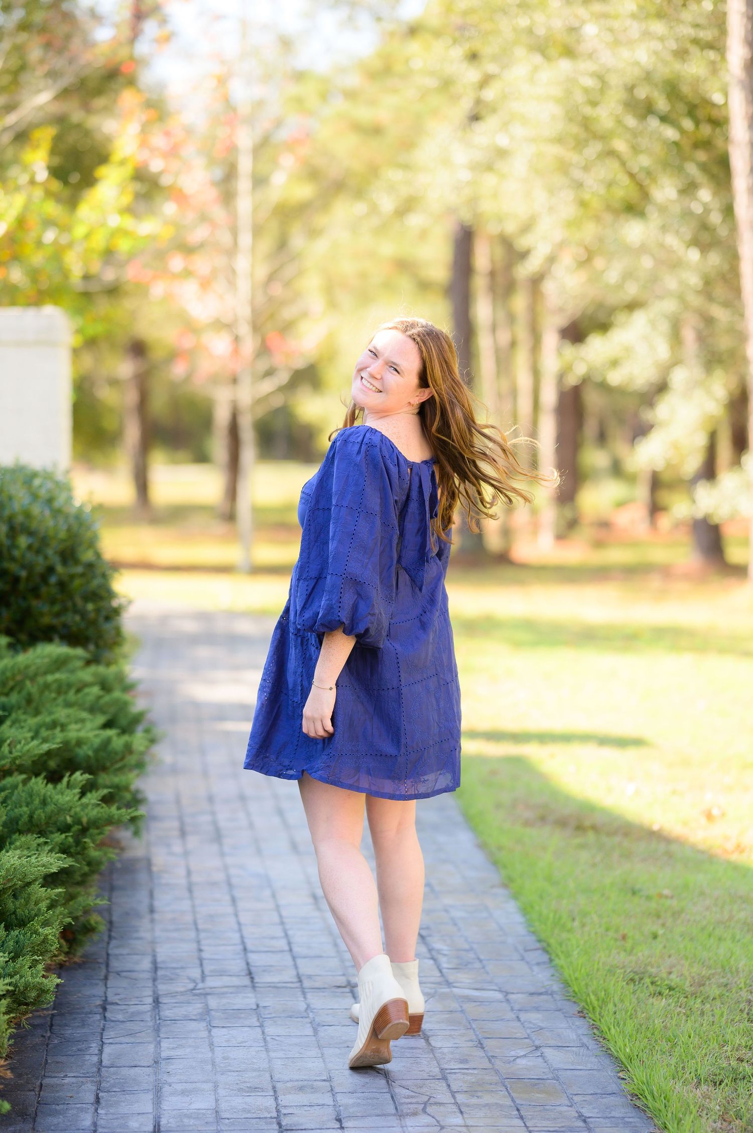 Woman in royal blue dress twirls playfully on a stone path surrounded by autumn trees.