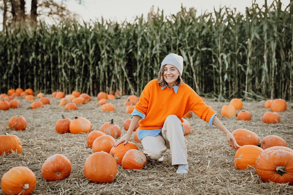 Person in orange sweater sits among pumpkins in front of a cornfield.