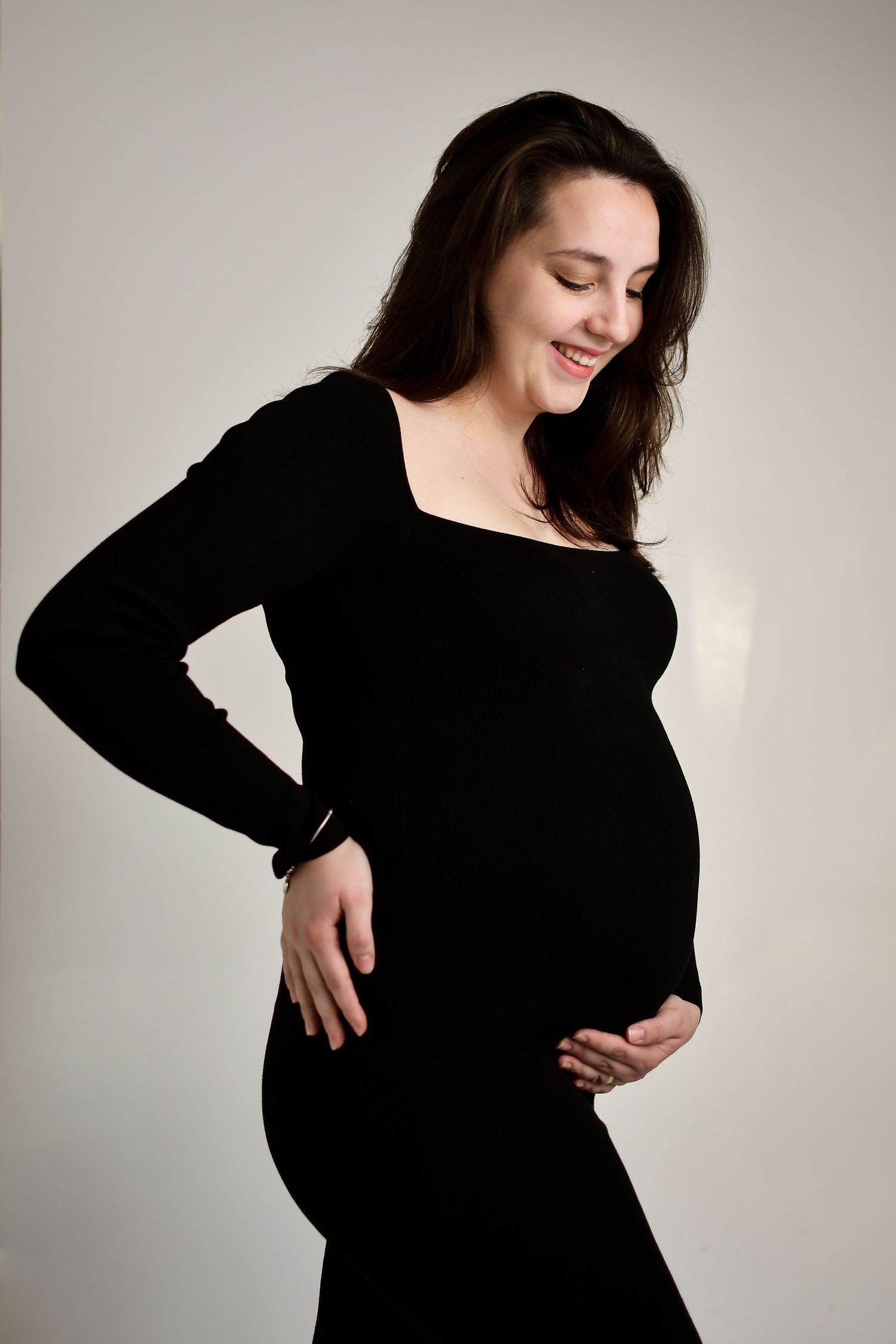 Studio maternity portrait of woman in black dress against light background.