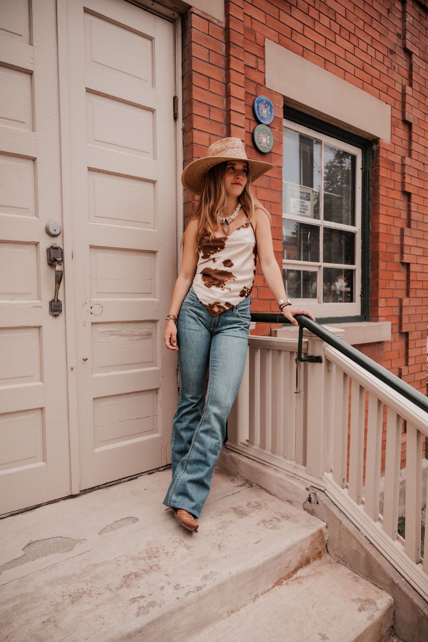 Summer western fashion shoot featuring a model in a brown cow print silk top and straw hat against a brick background.