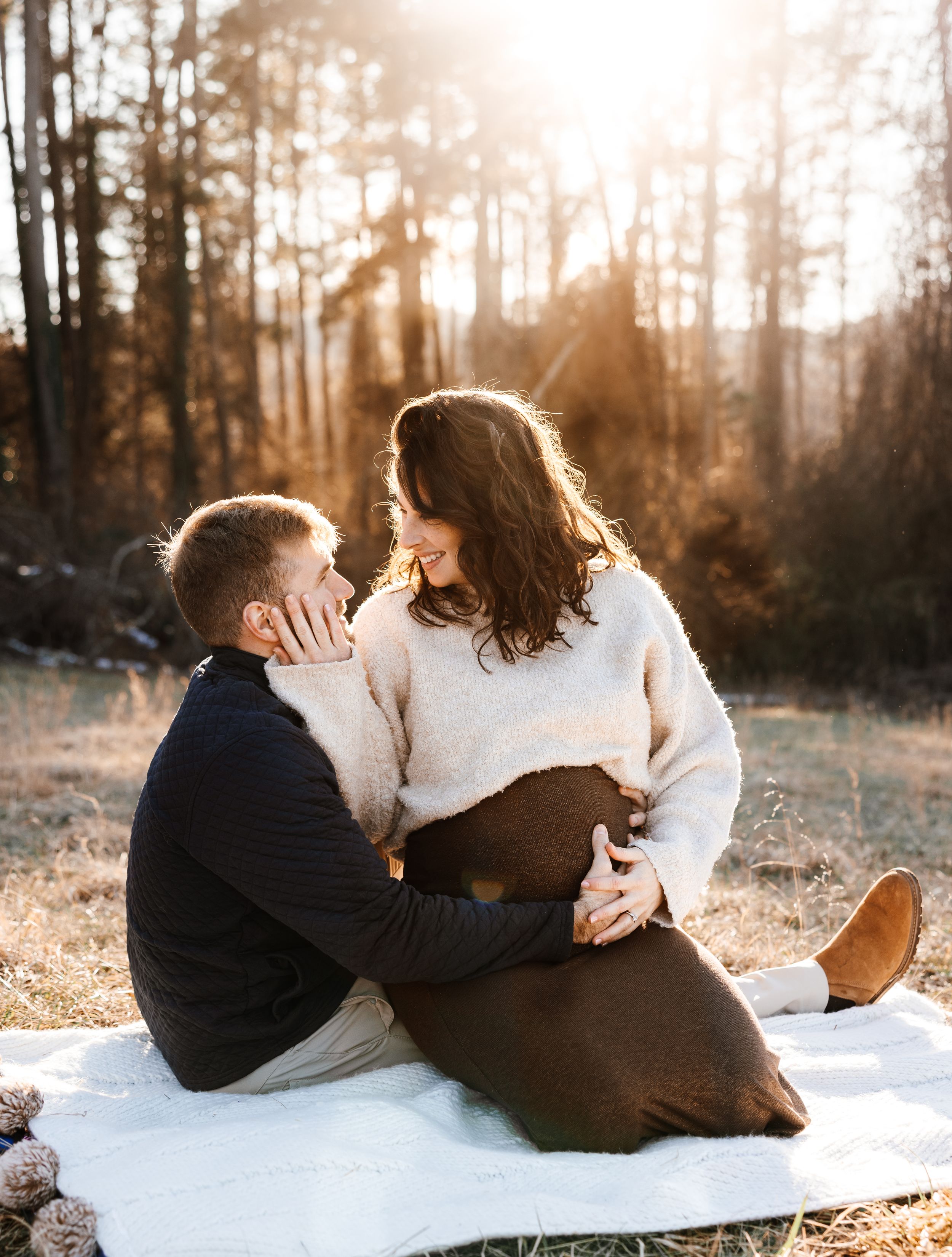 A romantic sunset engagement photo on a white blanket in a forest with warm golden light filtering through trees.