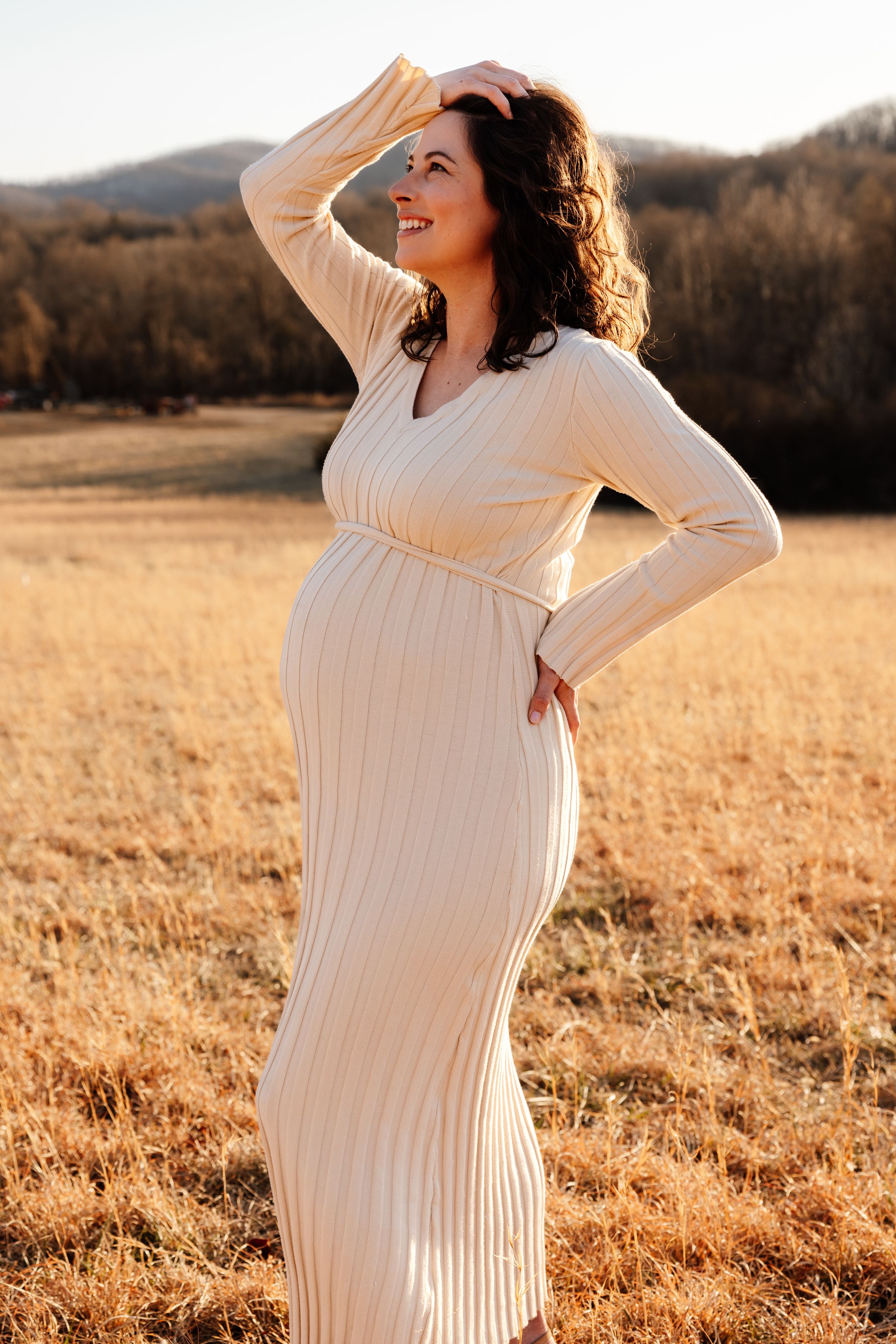 Expecting mother in white ribbed maternity dress poses gracefully in golden sunlit field at sunset while looking upward.