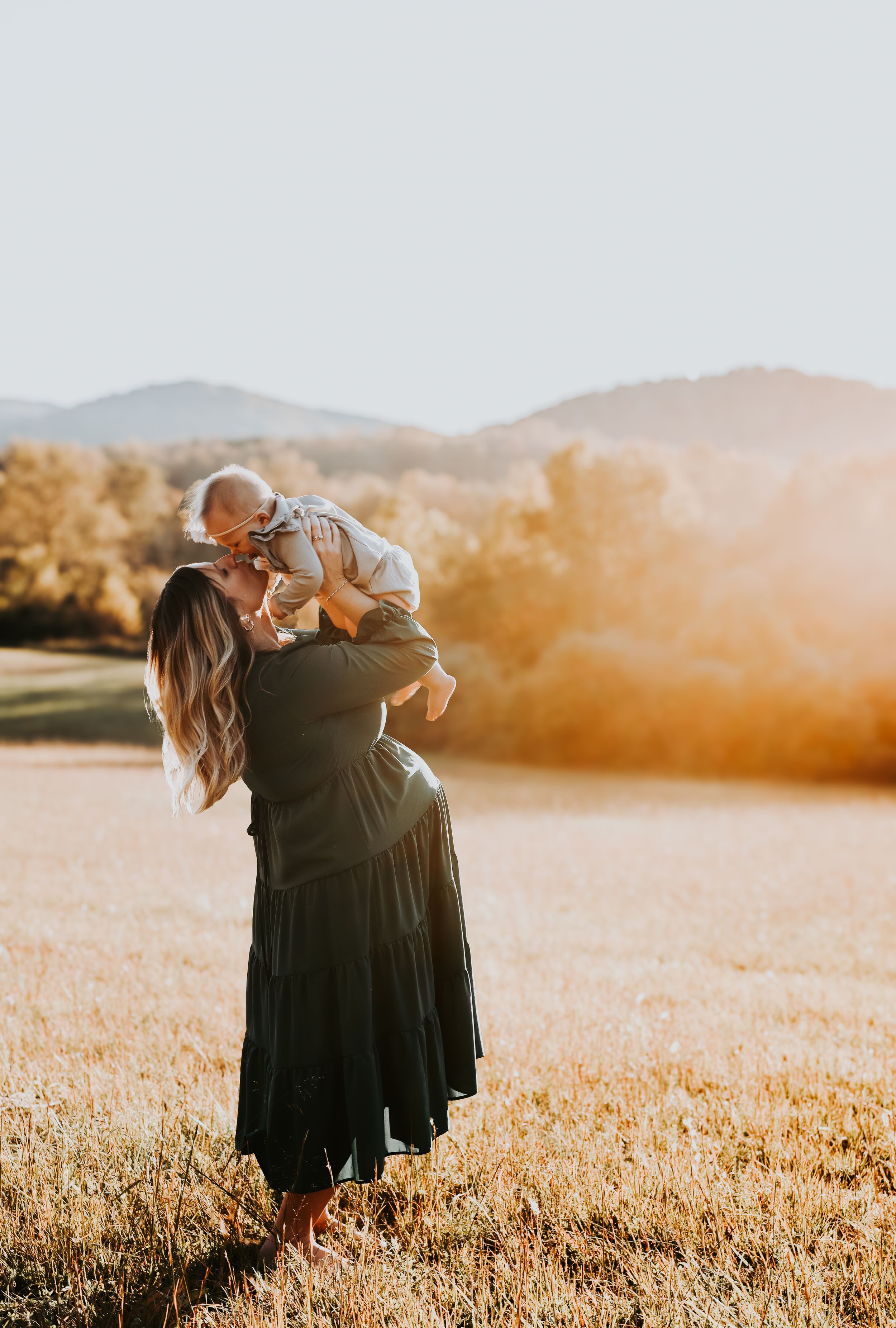 Mother lifting baby in golden field during sunset.