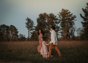 A couple walks together in a rustic field during sunset with pine trees in the background.