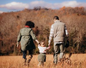 A family walking together through an autumn field with trees displaying orange and brown fall colors.
