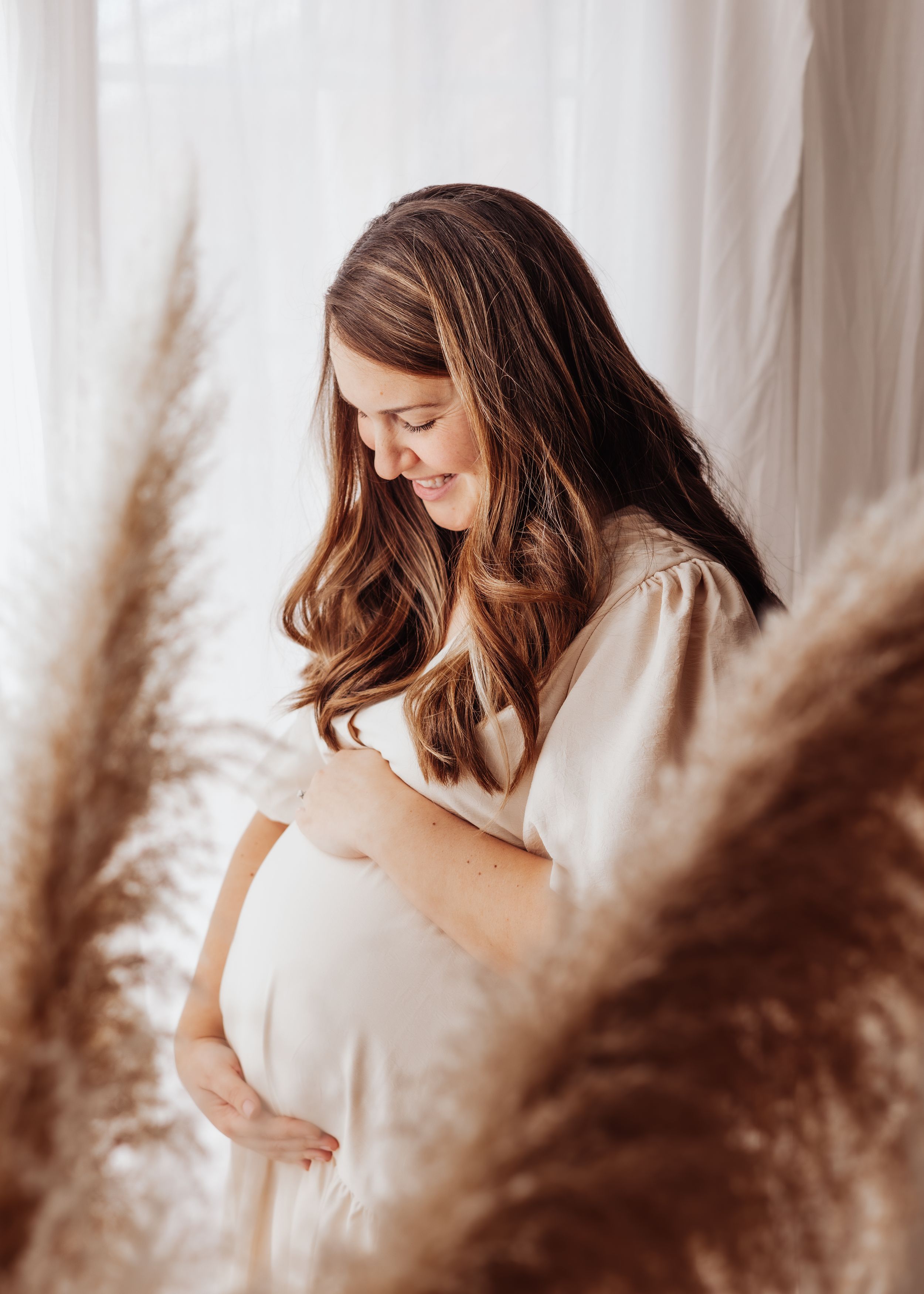 Expectant mother in white dress poses with pampas grass during maternity photoshoot.