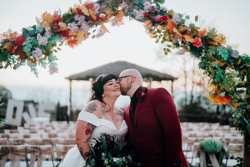 A couple shares a tender moment under a colorful floral arch at an outdoor wedding ceremony.
