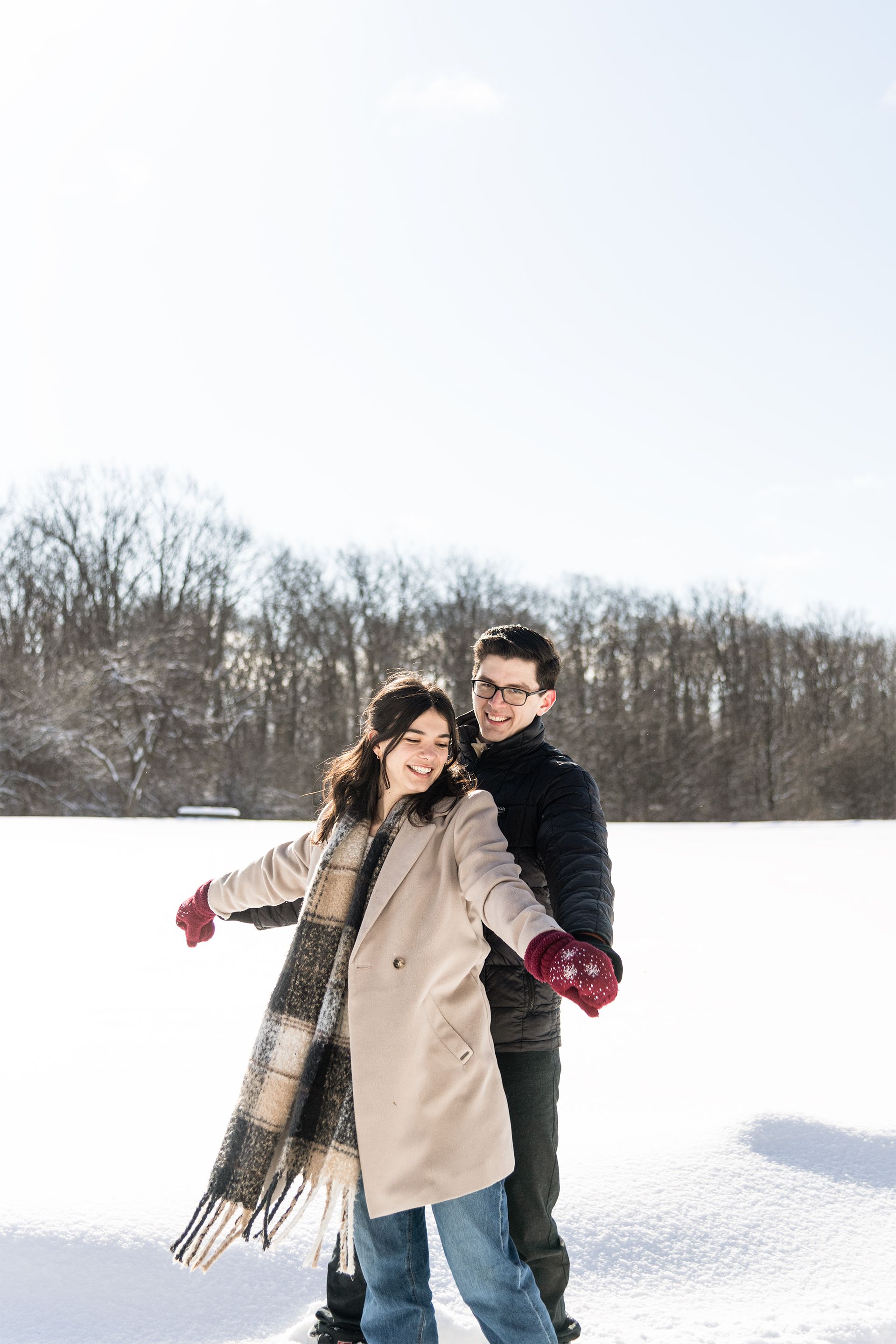 A couple wearing winter coats playfully poses together in a snowy field with bare trees in the background.