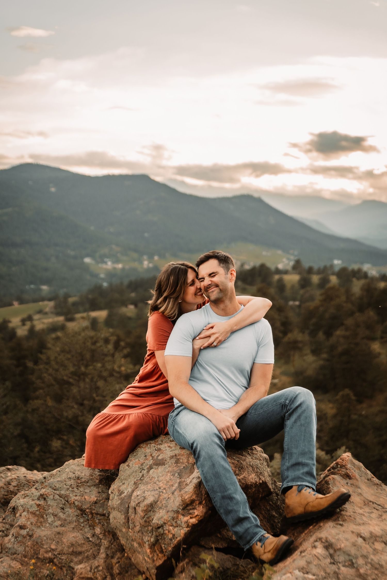 Couple shares intimate moment while sitting on rocky overlook with mountain vista at sunset.