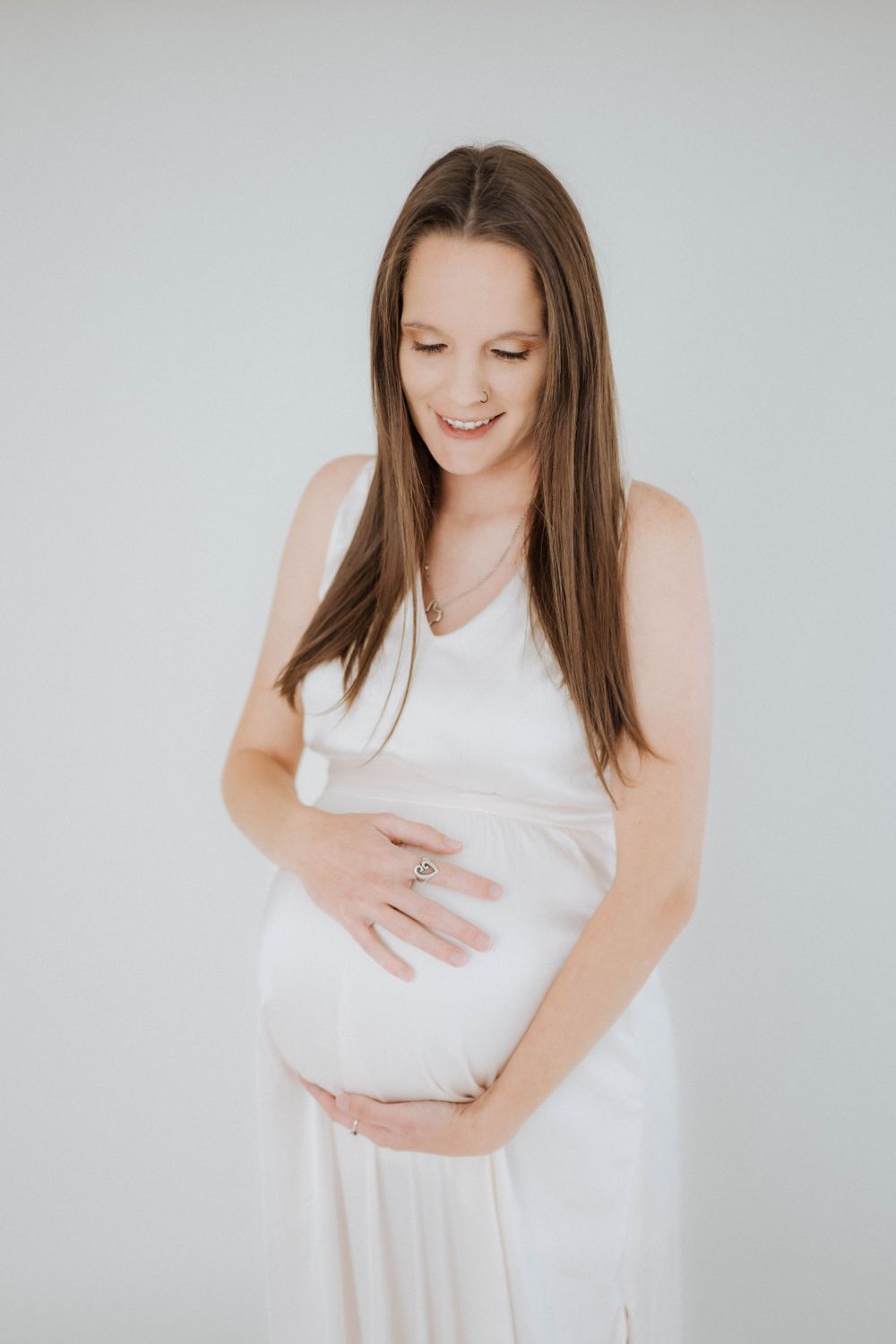 A pregnant woman in a white dress smiles while tenderly holding her baby bump against a light gray background.