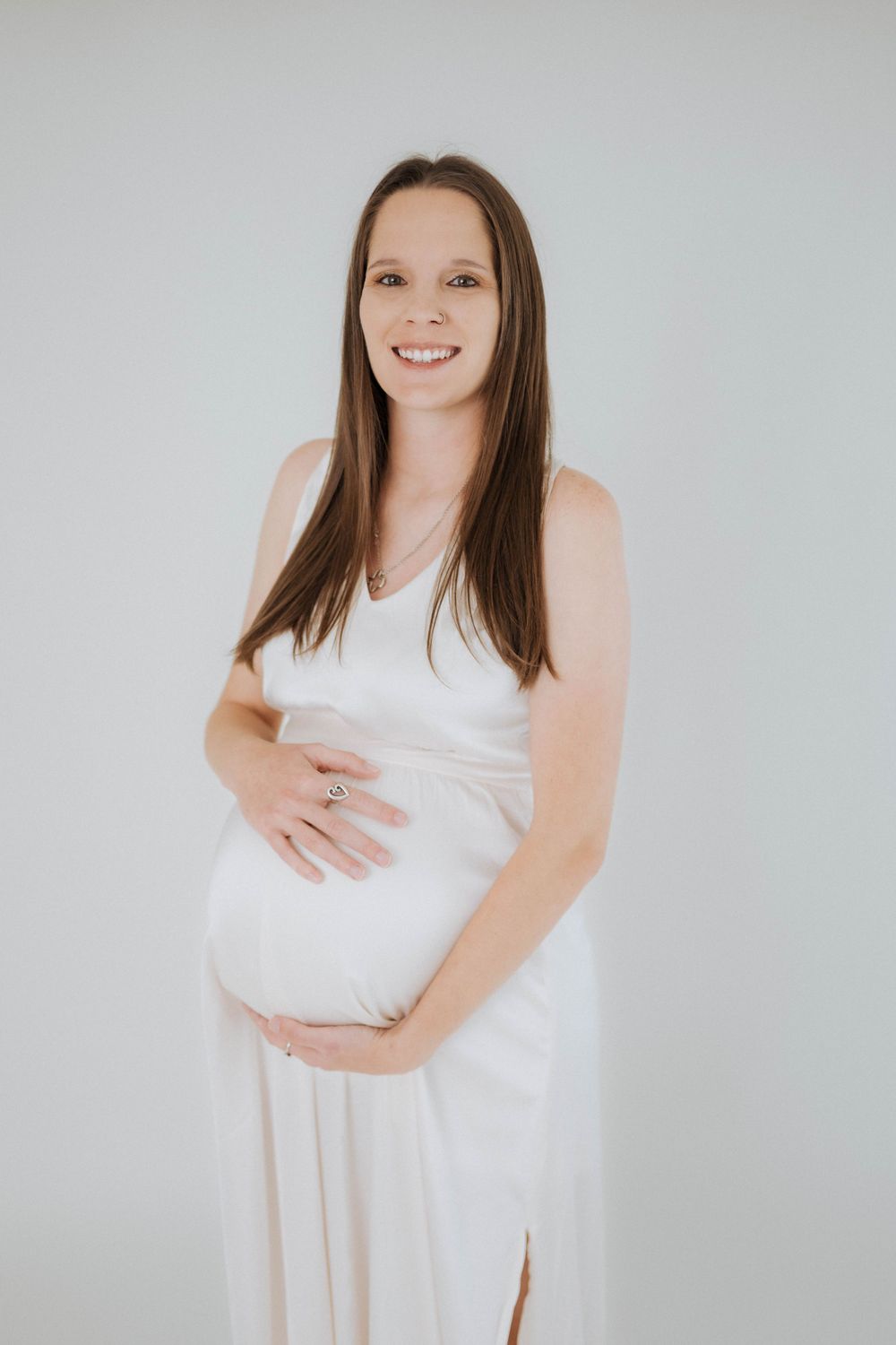 Expectant person wearing a white dress poses against grey background while cradling belly during maternity photoshoot.