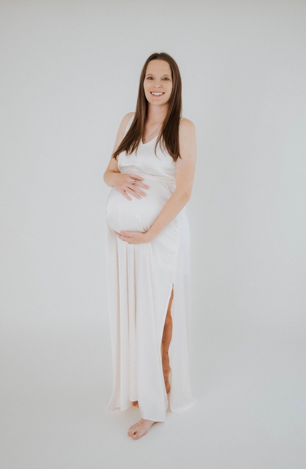 Expectant mother in flowing white maternity dress poses barefoot against light gray backdrop in studio setting.