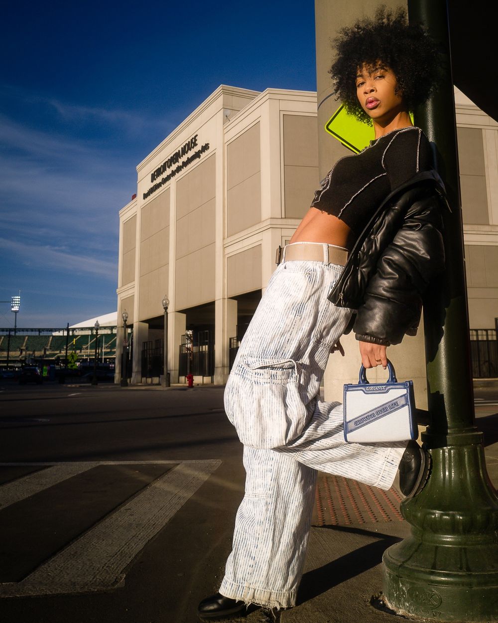 Fashion model poses against building exterior wearing black leather jacket, white cargo pants and neon yellow crop top.