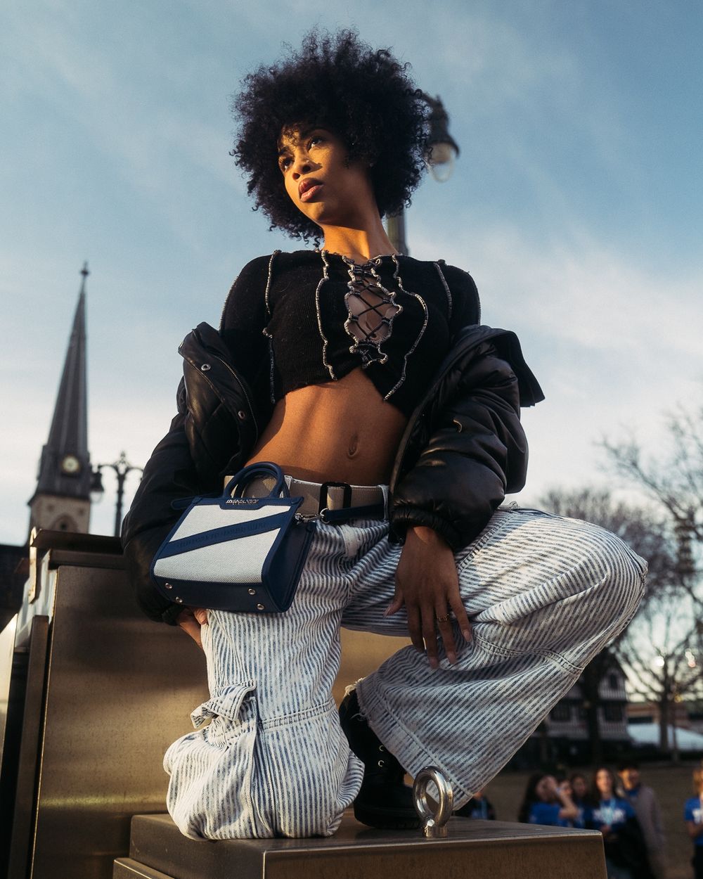 Street style fashion shot with black crop top, leather jacket and white striped pants against church spire.