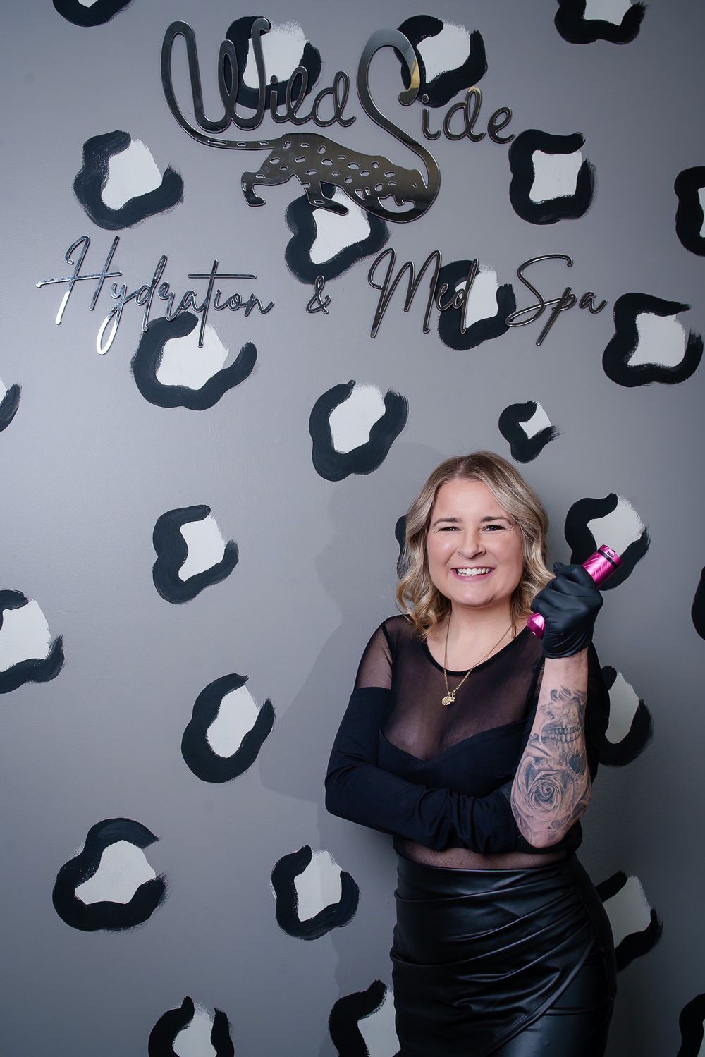 Salon professional in sheer black outfit poses against branded wall with leopard spot pattern.
