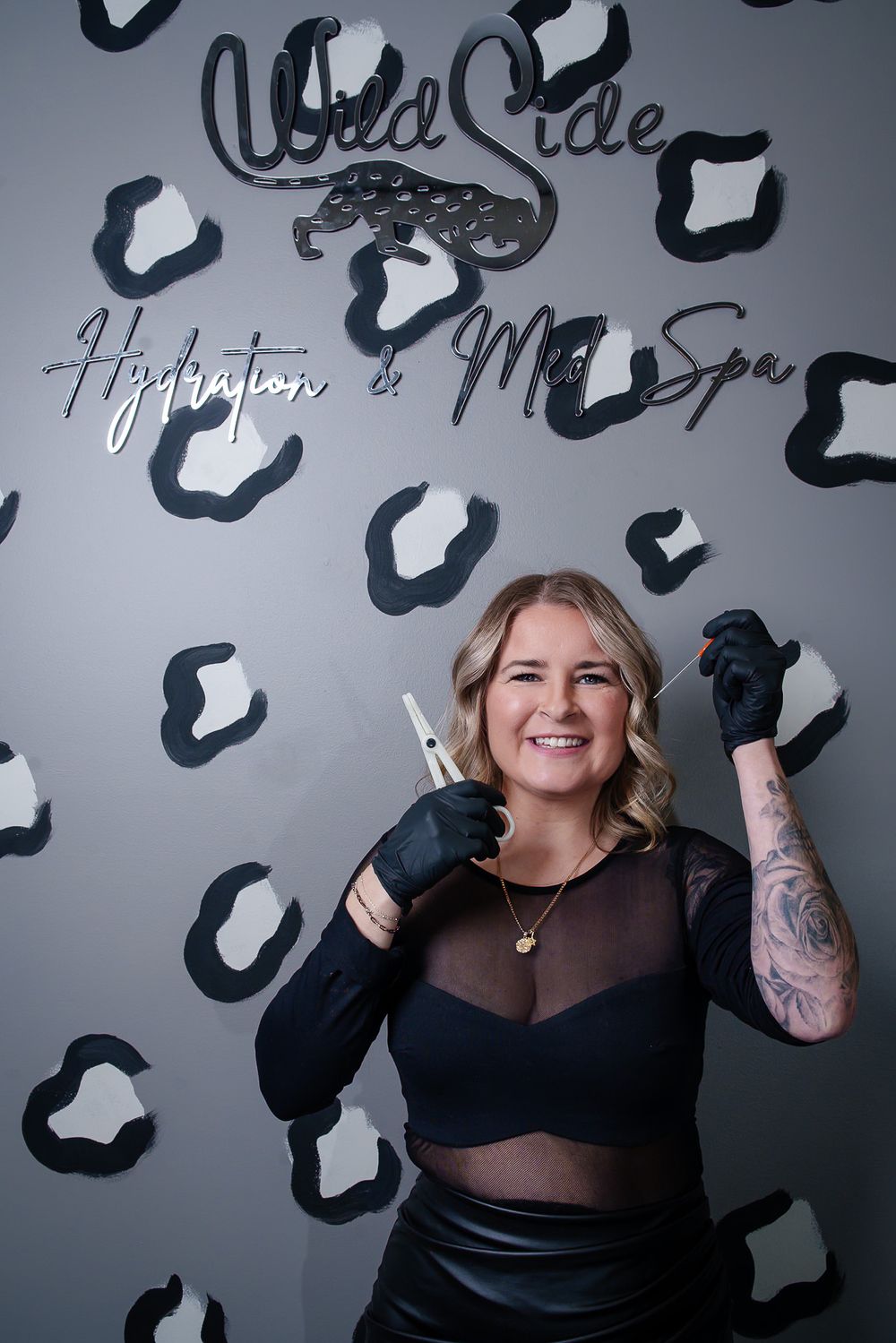 Stylist in sheer black outfit poses playfully against leopard spot wall backdrop.