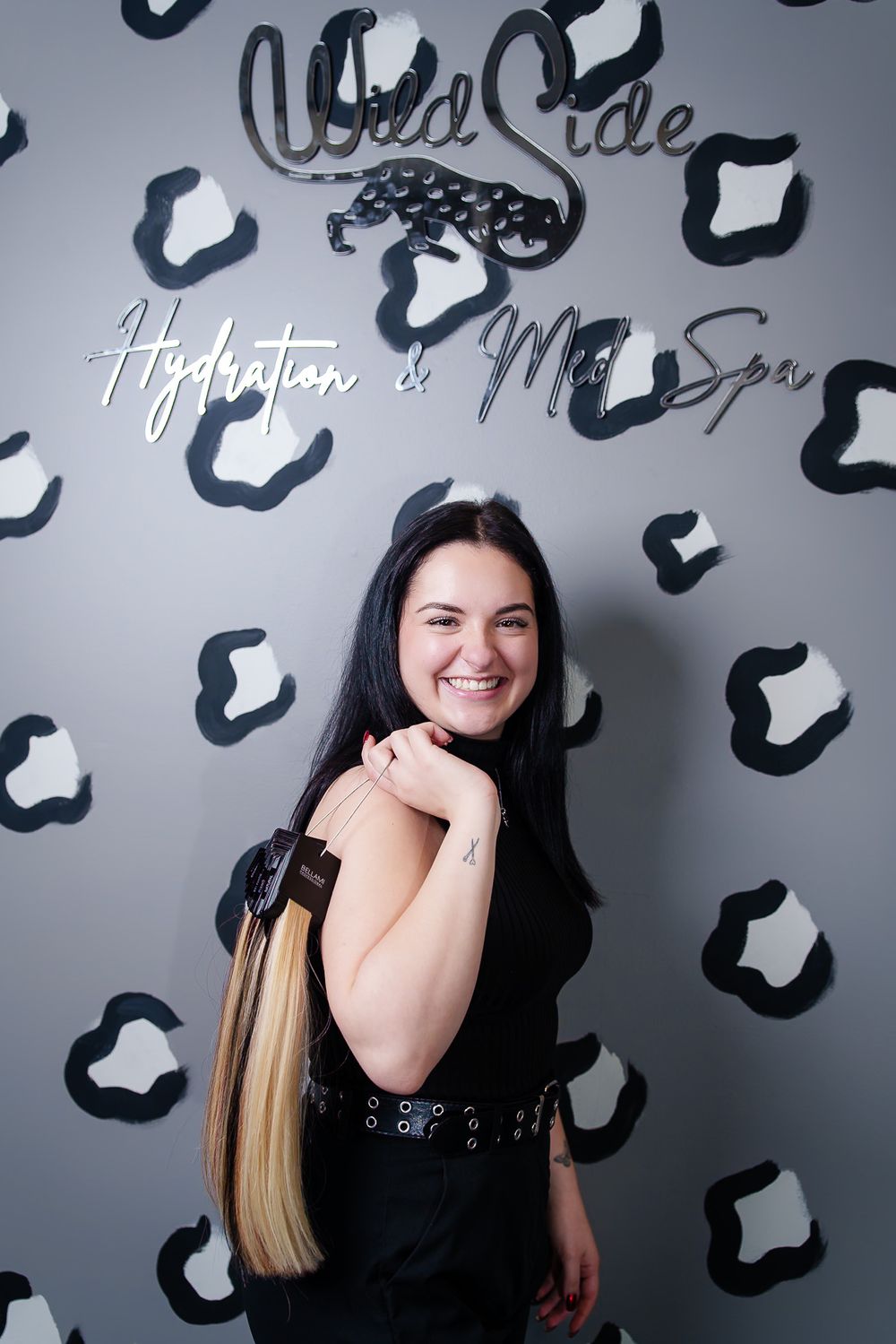 Hair stylist in black outfit poses against decorative salon wall with leopard print design.