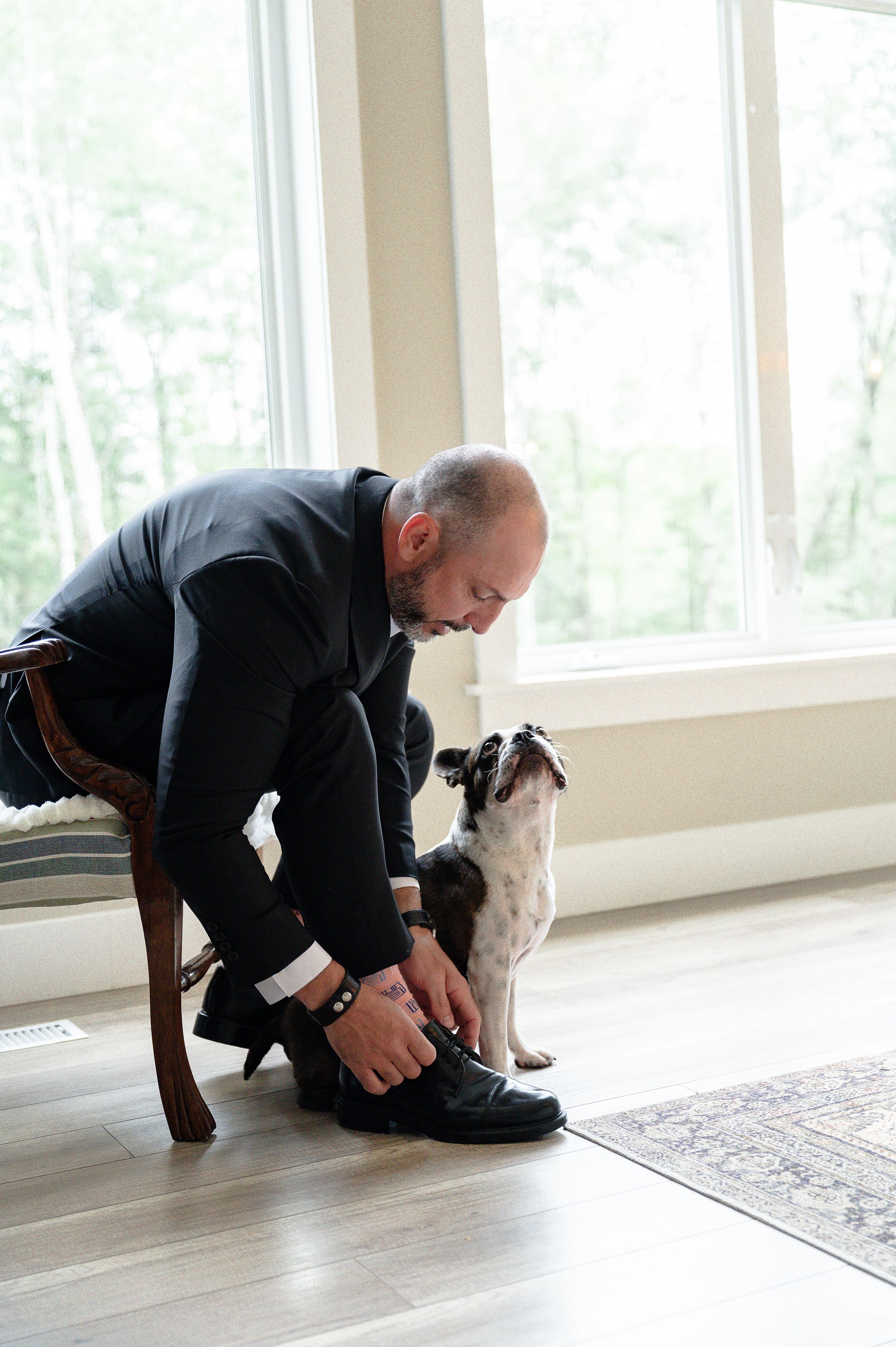 Groom tying his shoe while he gets ready for his wedding with his little French bulldog looking up at him.