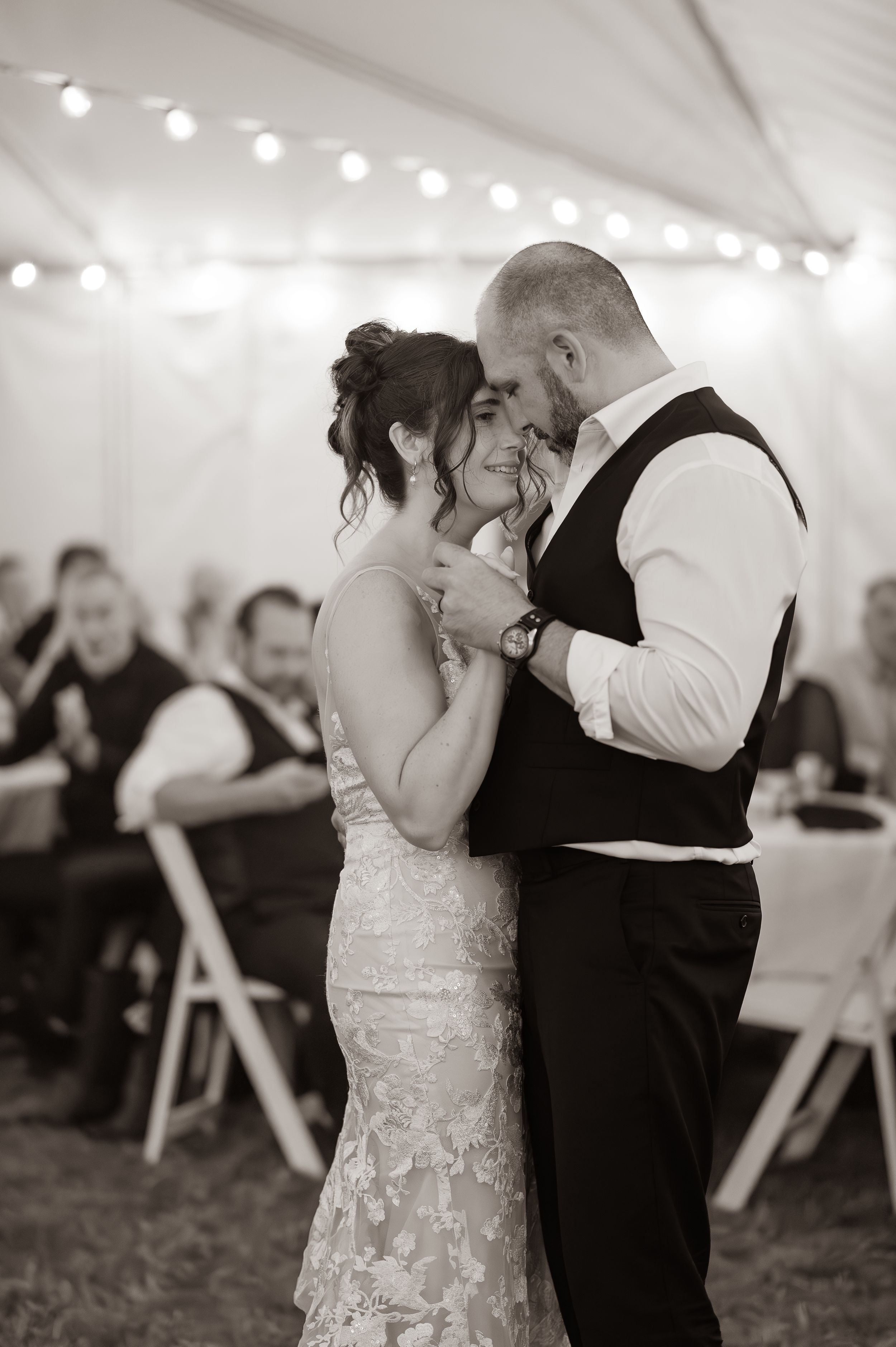 Newlyweds holding each other close and gazing into each other's eyes during their first dance with guests joyfully looking on in the back