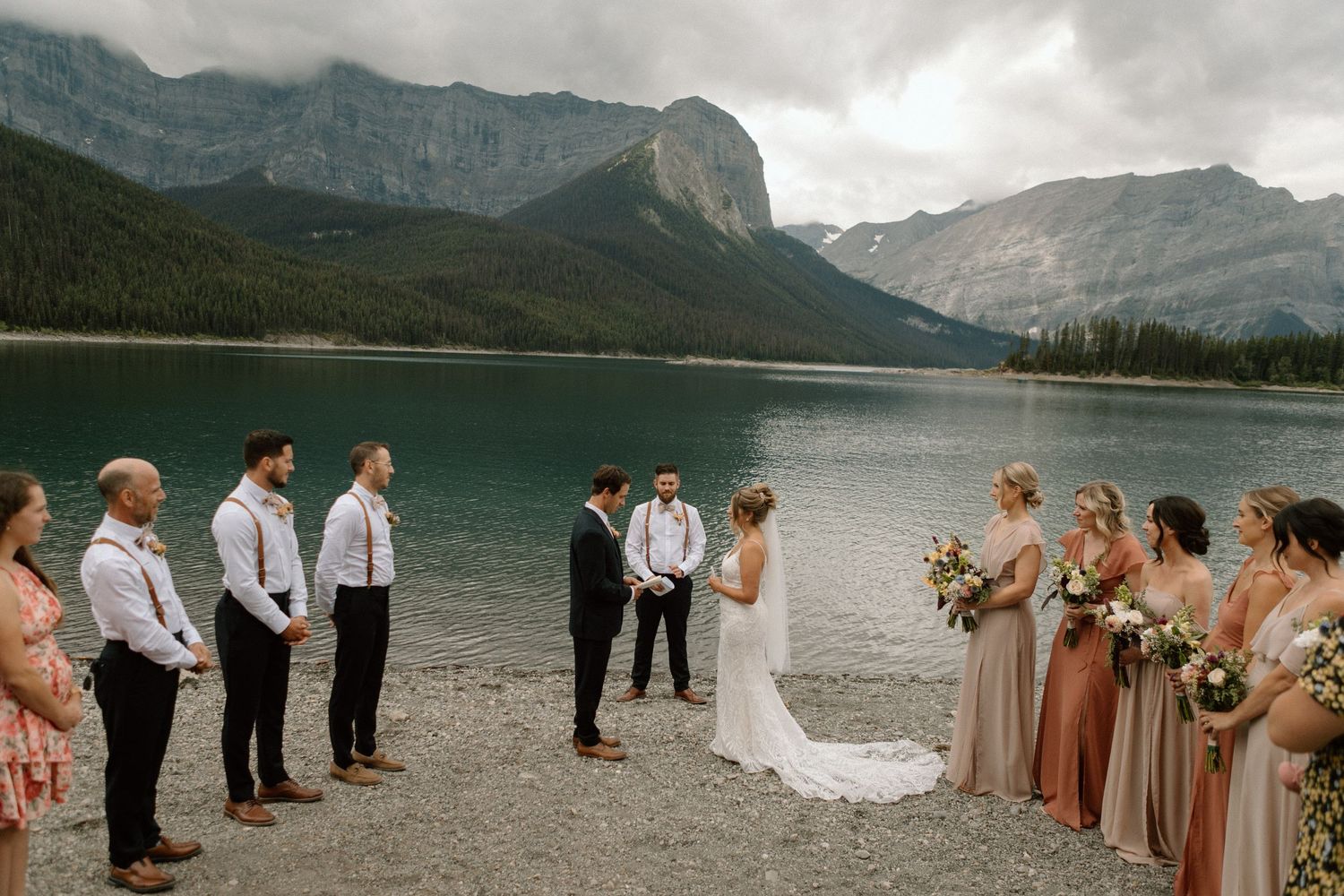 A wedding ceremony takes place on a rocky shore with dramatic mountain peaks and a lake in the background.