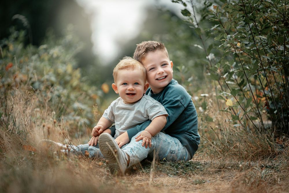 Two children sitting together outdoors in a natural setting with greenery and soft lighting in the background.