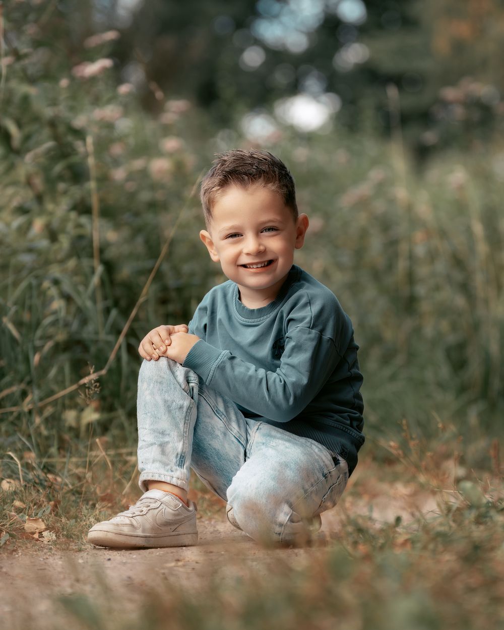 A child sits playfully in a field wearing a blue shirt and light-colored pants with white boots.