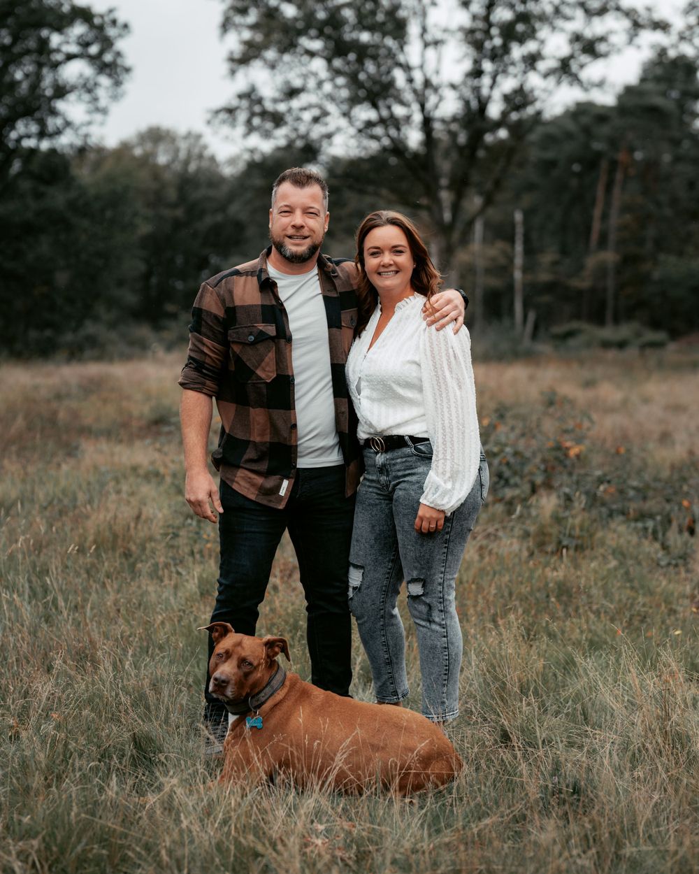 A couple poses with their brown dog in a rustic outdoor setting during autumn.
