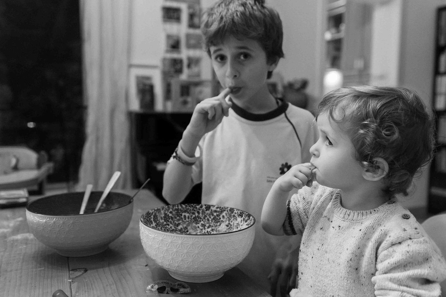 Two children eating from a large bowl at a kitchen table in a nostalgic black and white photograph.