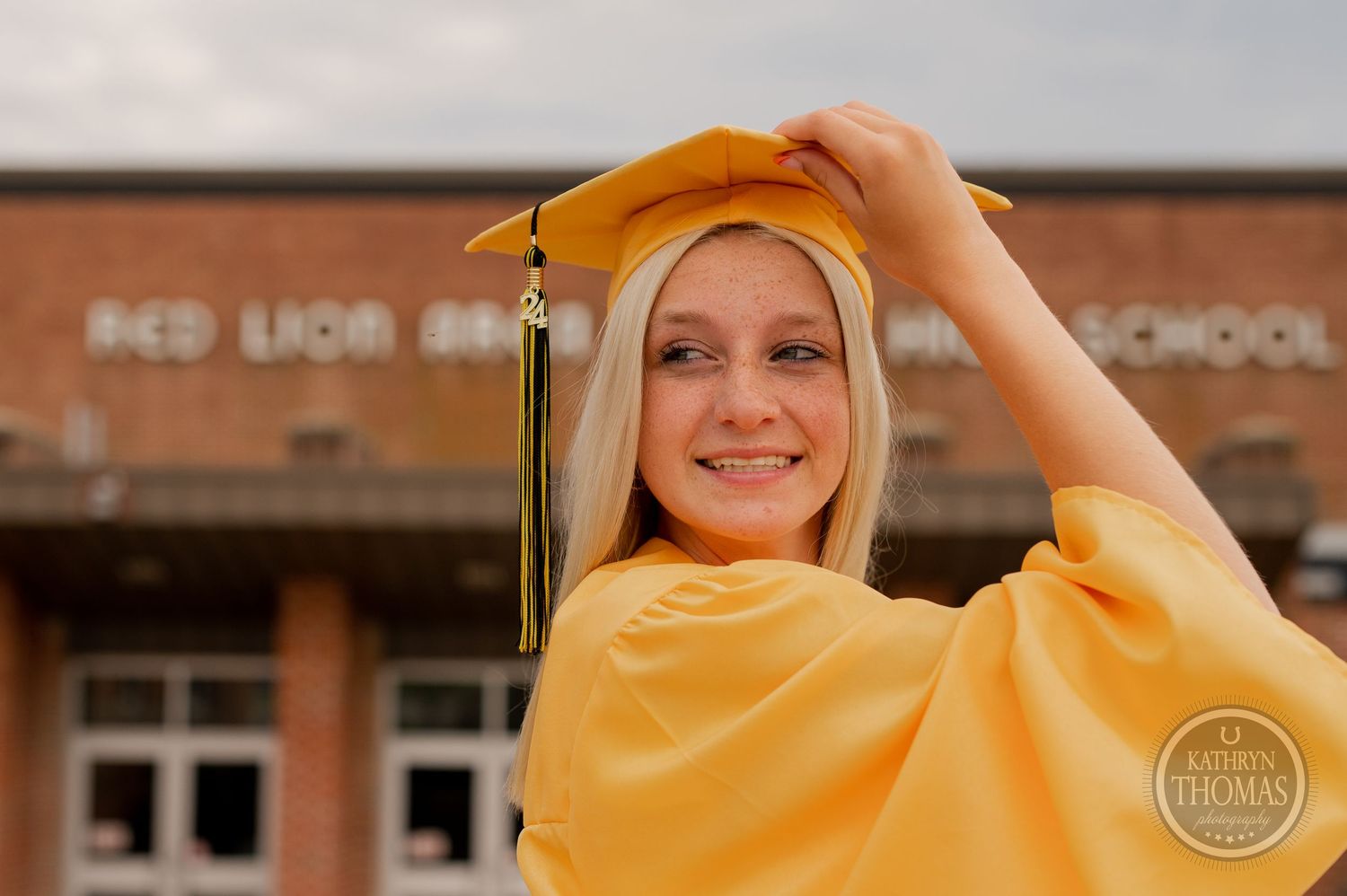 Graduate adjusts yellow cap and tassel while smiling in front of brick school building.