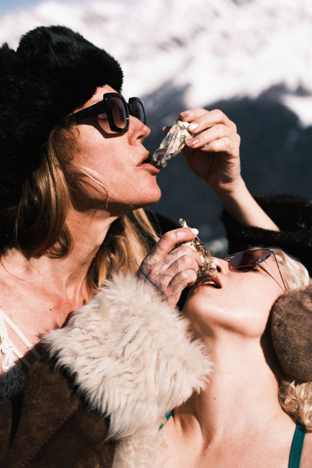 Two women in fur hats and sunglasses eating oysters outdoors with snowy mountains in the background.