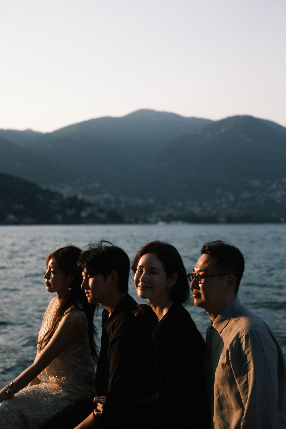 Four people standing by a lake with mountain backdrop at dusk, captured in a moody side profile portrait.