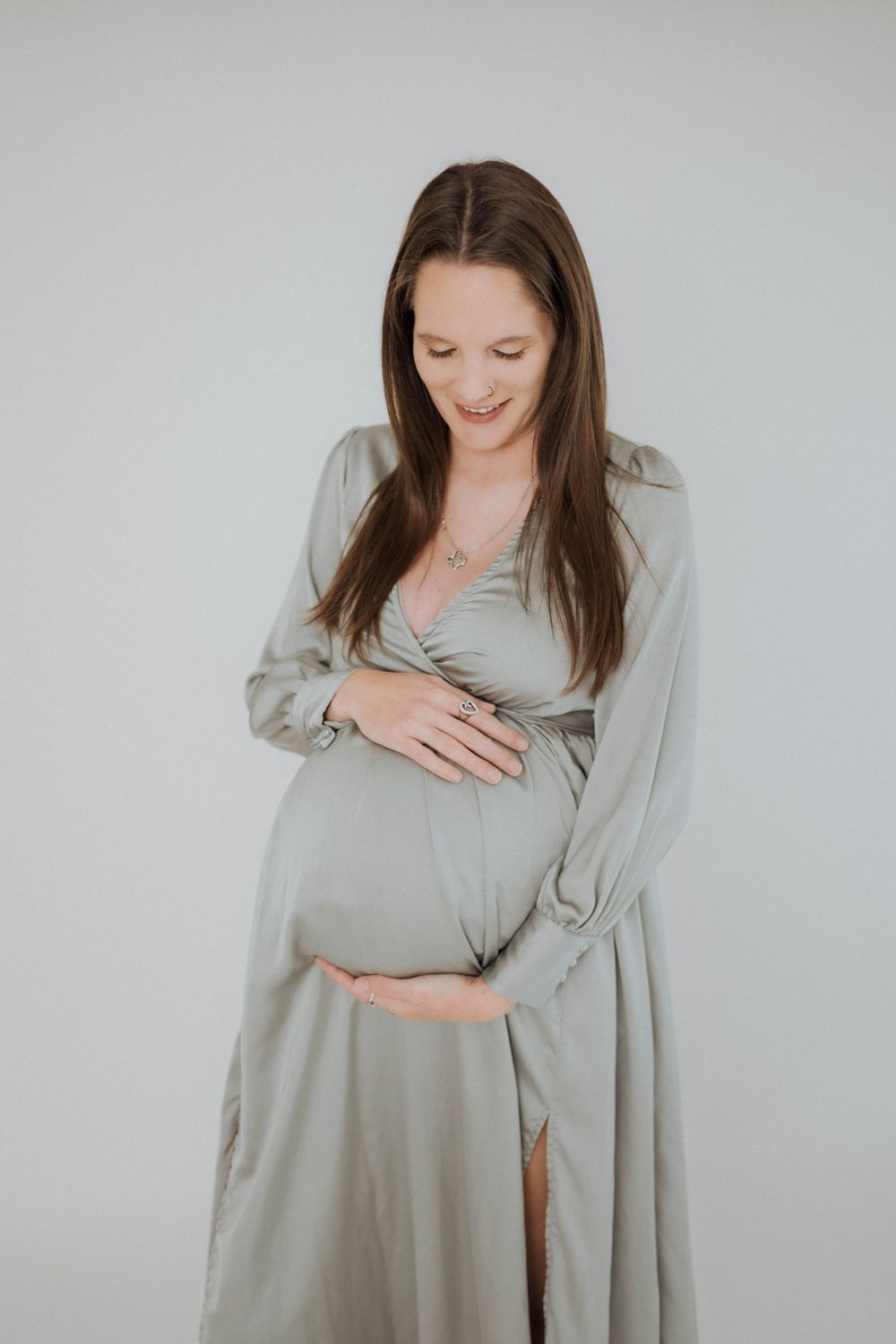 An expectant mother in a light gray flowing dress gently cradles her baby bump against a neutral background.