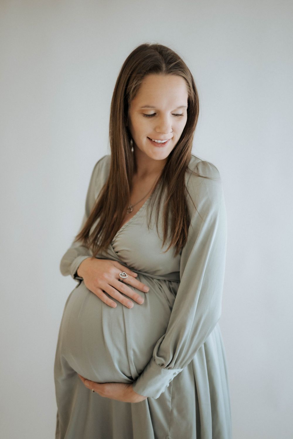 Expectant person in a flowing sage green maternity dress smiles while gently cradling their baby bump against a white backdrop.