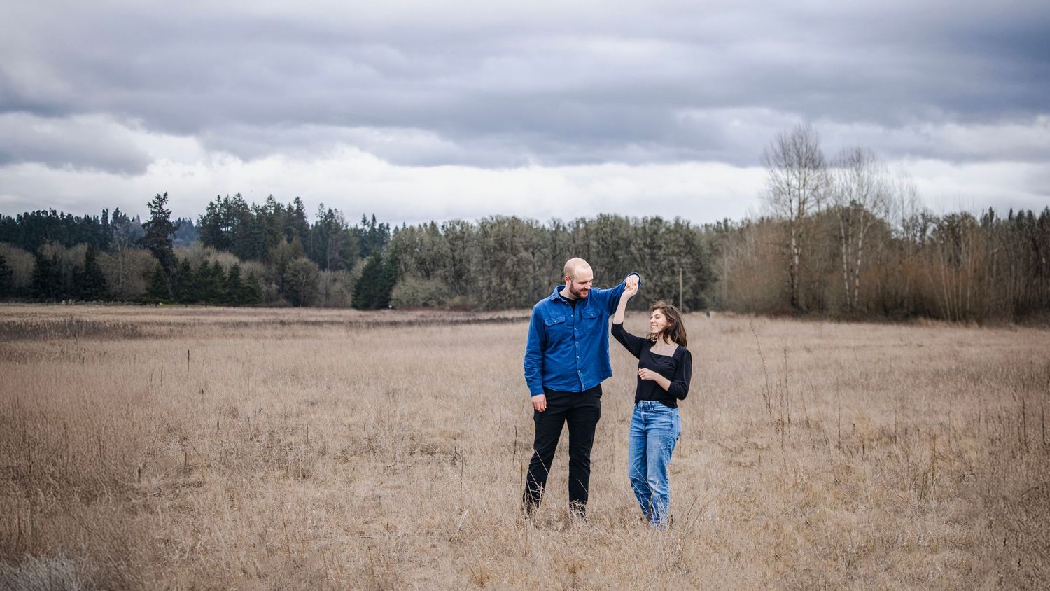 a man spins a woman in a large open field as they smile at each other. the field is golden, the sky is moody