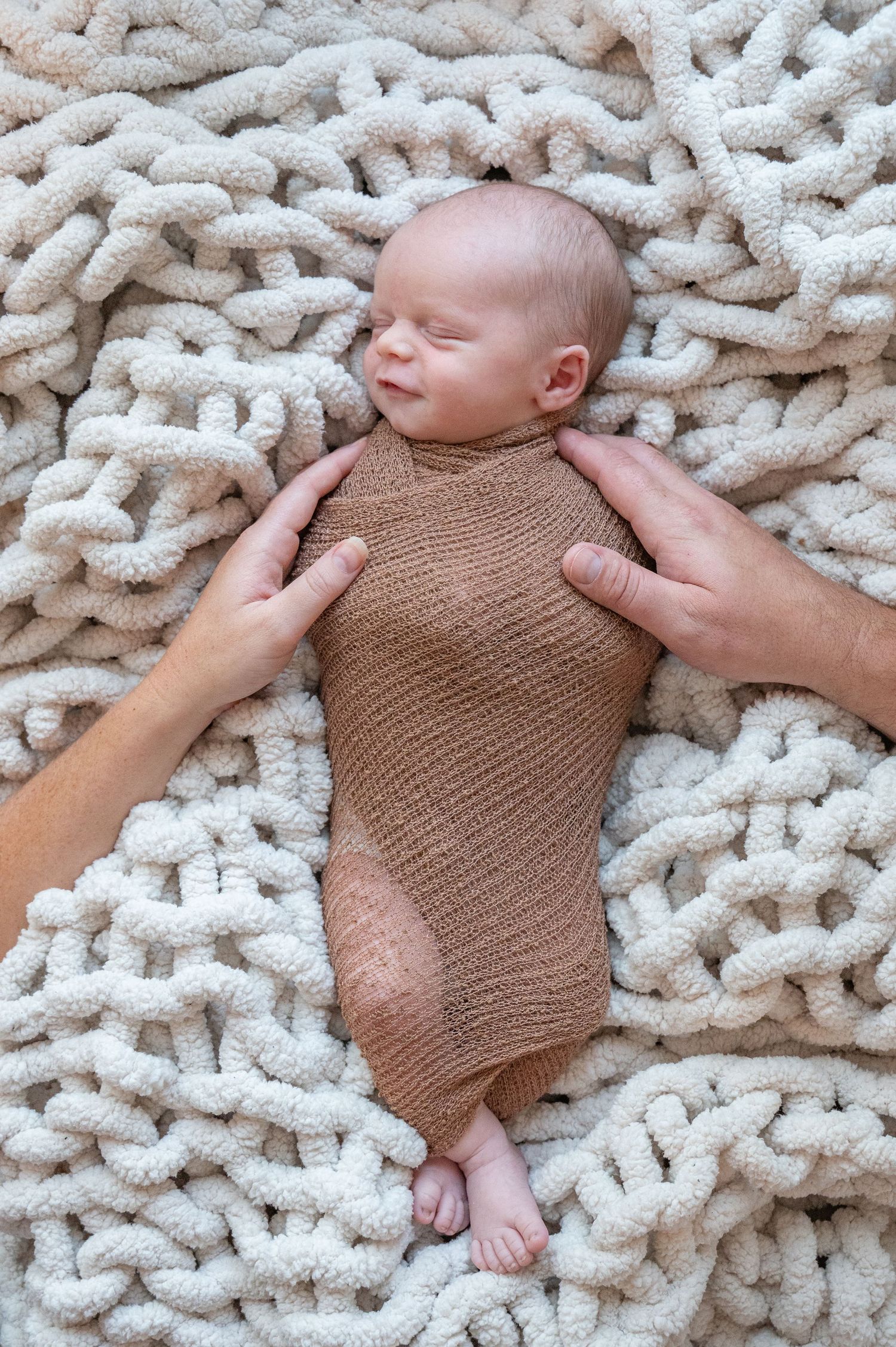 Newborn baby wrapped in brown knit fabric lying on chunky white knitted blanket.
