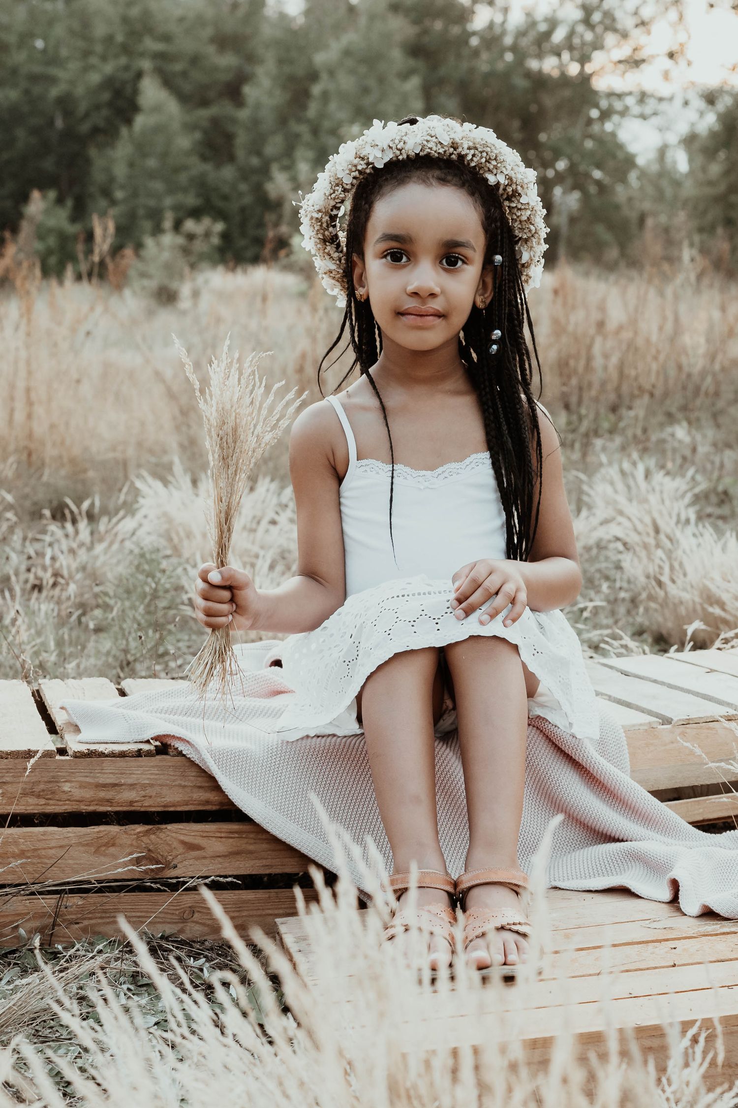 Person in white dress with flower crown sitting on wooden platform among tall grass.