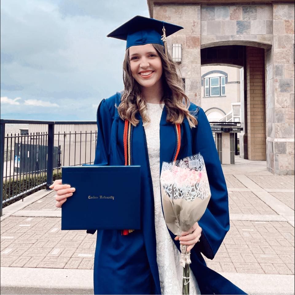 Jess celebrating graduation while holding diploma and bouquet in front of stone building