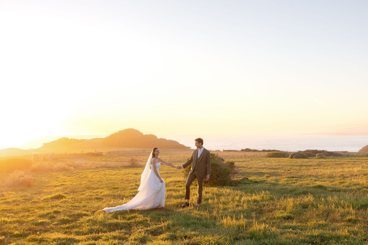 Couple in wedding attire holding hands in golden field at sunset with mountains in background.