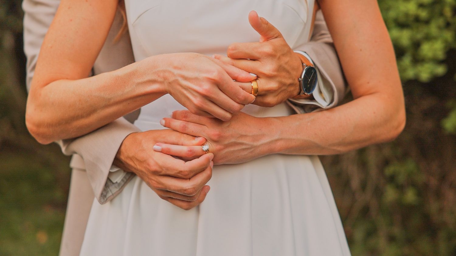 Close up view of hands holding each other wearing wedding rings against a white dress in an outdoor setting.