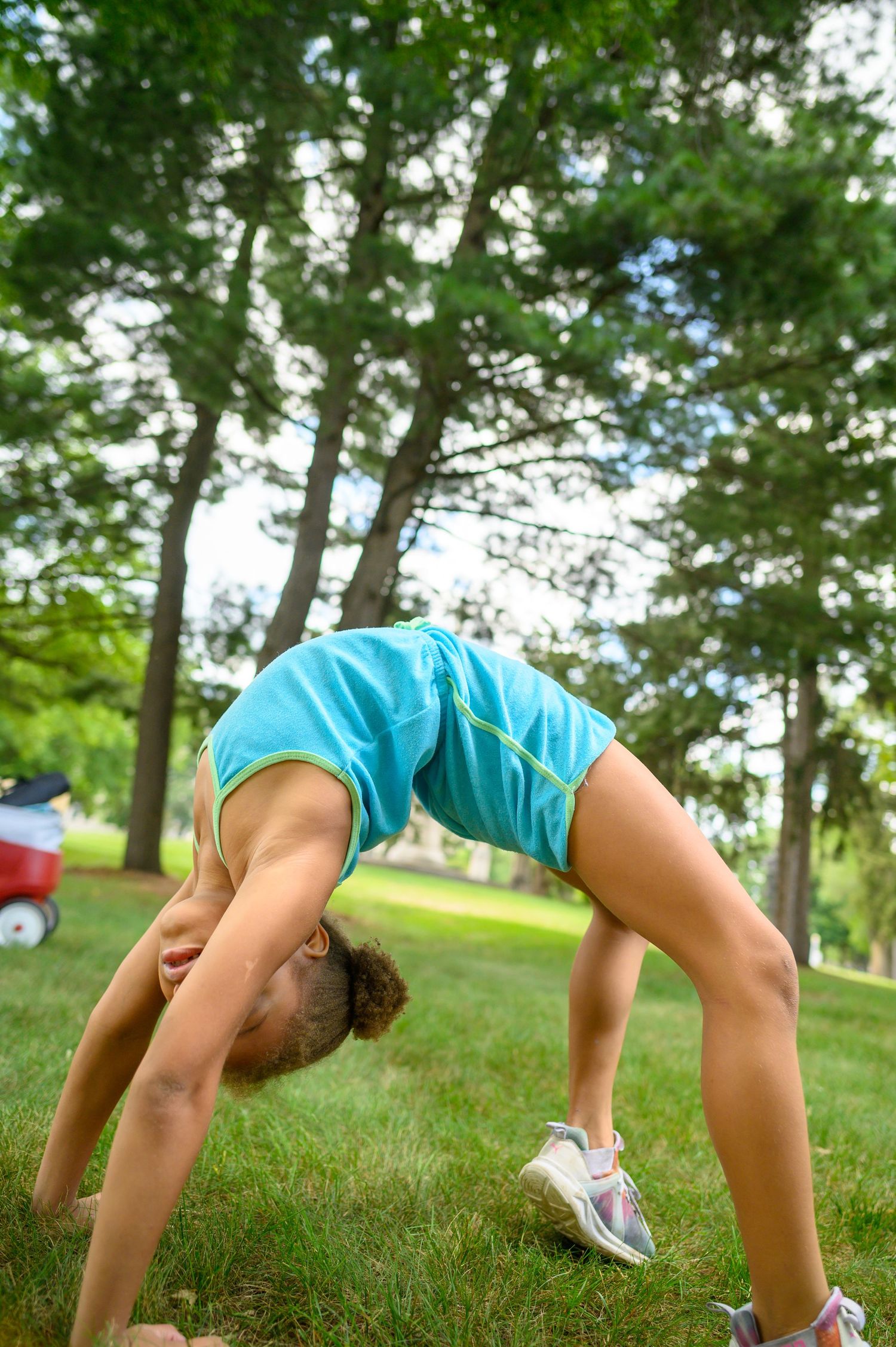 A gymnast performs a backbend in a turquoise outfit on grass with trees in the background.