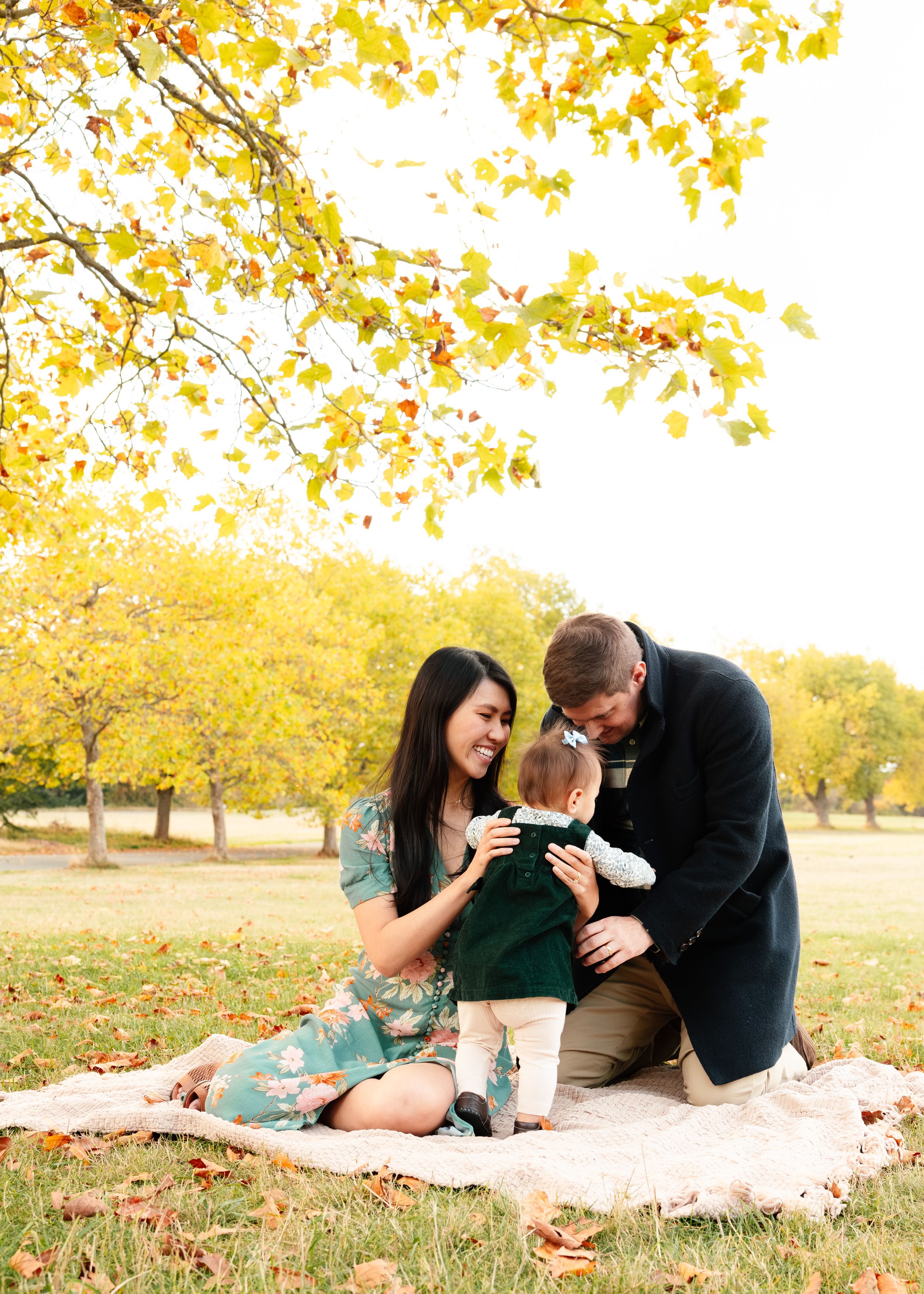 A couple sits on a blanket under golden autumn trees with yellow leaves playing with their baby.